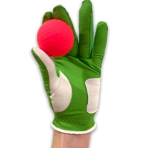 Op de afbeelding: Een felgroene golfhandschoen met witte accenten houdt een felroze golfbal vast. De handschoen heeft een textuur en een witte rand. De golfbal heeft een opvallende kleur.