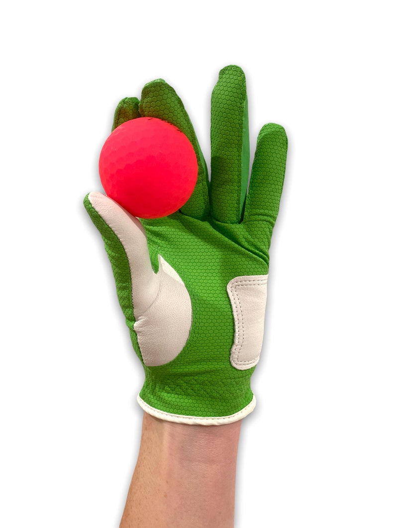 Op de afbeelding: Een felgroene golfhandschoen met witte accenten houdt een felroze golfbal vast. De handschoen heeft een textuur en een witte rand. De golfbal heeft een opvallende kleur.