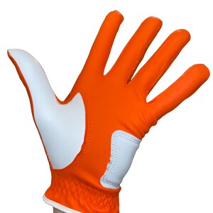 Op de afbeelding: Oranje en witte golfhandschoen. De handschoen is voornamelijk oranje met witte accenten op de handpalm en duim. De pols heeft een elastische band en witte rand. Ontworpen voor rechtshandig gebruik.