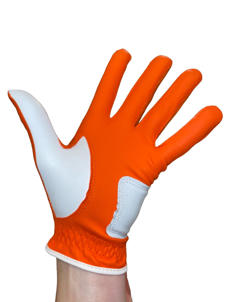 Op de afbeelding: Oranje en witte golfhandschoen. De handschoen is voornamelijk oranje met witte accenten op de handpalm en duim. De pols heeft een elastische band en witte rand. Ontworpen voor rechtshandig gebruik.