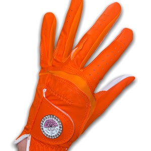 Op de afbeelding: Fel oranje golfhandschoen met witte rand en een decoratief embleem. De handschoen heeft een ademend ontwerp met kleine perforaties en een wit accent op de pols. Het embleem heeft een roze bloemenontwerp en de woorden "Love, Sue".