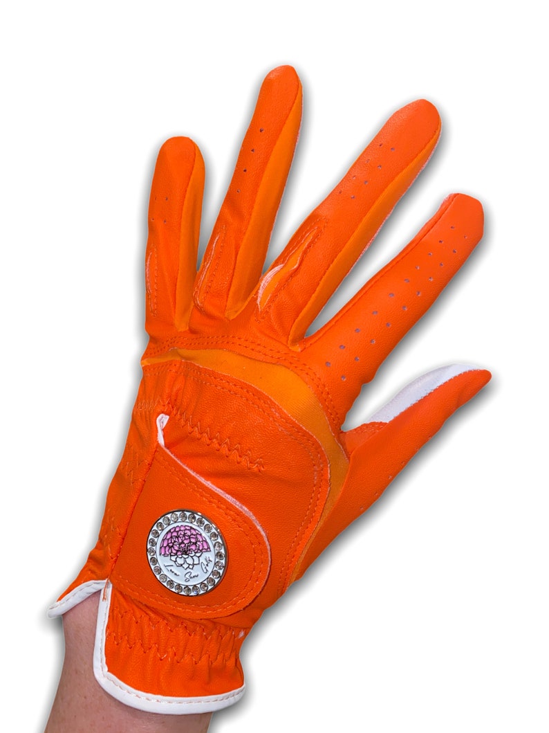 Op de afbeelding: Fel oranje golfhandschoen met witte rand en een decoratief embleem. De handschoen heeft een ademend ontwerp met kleine perforaties en een wit accent op de pols. Het embleem heeft een roze bloemenontwerp en de woorden "Love, Sue".