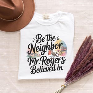 May include: White t-shirt featuring the phrase "Be the Neighbor Mr. Rogers Believed in." The design includes a bird, houses, and a tram. A brown hat is visible in the upper left corner. The shirt is folded.