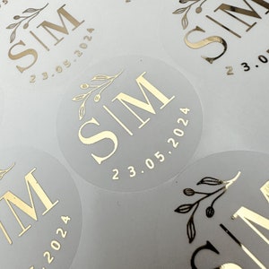 May include: A collection of circular translucent stickers featuring gold foil lettering. Each sticker displays the letters SIM in a serif font, a small botanical sprig illustration above the text, and the date 23.05.2024 printed in gold.
