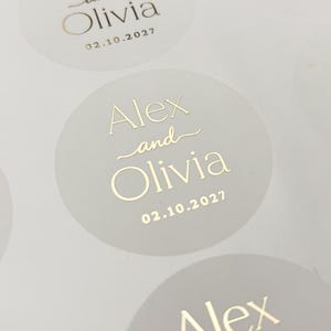 May include: A sheet of circular stickers featuring gold foil text that reads Alex and Olivia with the date 02.10.2027 printed below. The stickers are arranged in a grid pattern on a white backing sheet, showcasing a clean and uniform design.
