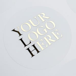 Könnte beinhalten: Transparenter, runder Aufkleber mit goldfarbenem Schriftzug "YOUR LOGO HERE". Der Text ist kreisförmig angeordnet. Der Aufkleber befindet sich auf weißem Hintergrund. Ideal für Branding und Produktkennzeichnung.