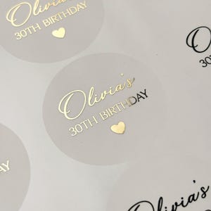May include: Round, semi-transparent stickers featuring gold foil text that reads Olivia's 30TH BIRTHDAY, accompanied by a small gold heart icon. The design is printed on a matte, light-coloured circular label sheet, showcasing a clean and elegant aesthetic.