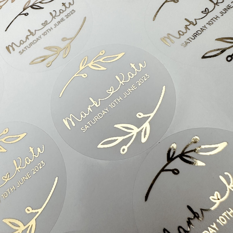 May include: Clear round stickers with gold foil lettering and a simple branch design. The text reads "Mark & Kate" and "Saturday 10th June 2023".