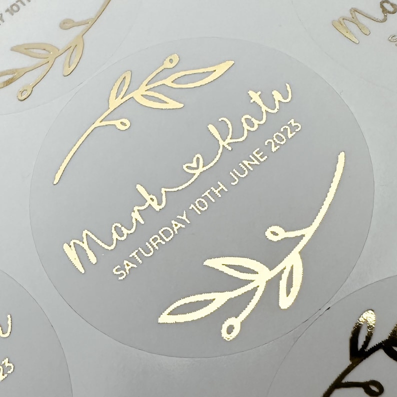May include: Round, white sticker with gold foil lettering and a simple gold leaf design. The sticker says "Mark & Kat" and "Saturday 10th June 2023".