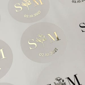 May include: Round, light beige stickers featuring a gold foil design with the letters S and M separated by a floral illustration. Below the letters, the date 02.10.2027 is printed in gold foil. The stickers are arranged on a white backing sheet.