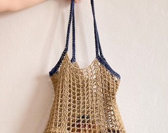 May include: A beige crocheted tote bag with navy blue straps and trim. The open weave design reveals the contents inside. The bag is held up by a hand against a white background. The bag is a stylish and eco-friendly accessory.