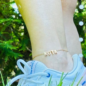 May include: A gold ankle bracelet with the name "KOTA" spelled out in block letters. The bracelet is worn on a person's ankle above a white trainer.