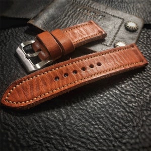 Peut inclure: Un bracelet de montre en cuir marron avec une boucle argentée. Le bracelet a un bord cousu et une surface texturée.