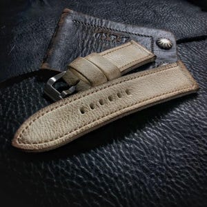 May include: A light beige leather watch strap with stitching around the edges. The strap has a silver buckle and is shown on a dark brown leather surface.