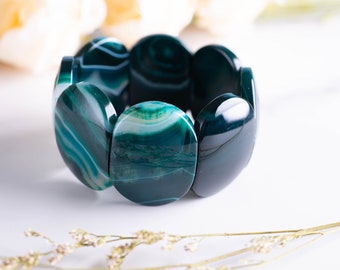 May include: A stretch bracelet featuring large, oval-shaped beads. The beads are a deep teal color with white and teal striations. The bracelet is displayed on a white surface with soft, blurred floral accents in the background.