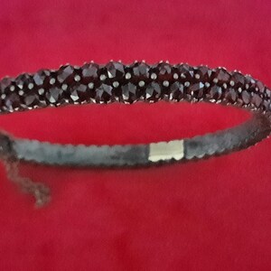 K&ouml;nnte beinhalten: Ein silbernes Armband mit einer Reihe dunkelroter Edelsteine. Das Armband ist offen und hat einen Verschluss.