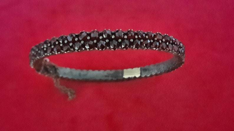 K&ouml;nnte beinhalten: Ein silbernes Armband mit einer Reihe dunkelroter Edelsteine. Das Armband ist offen und hat einen Verschluss.