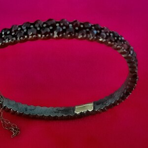 K&ouml;nnte beinhalten: Ein silbernes Armband mit einer Reihe dunkelroter Edelsteine. Das Armband hat einen Verschluss an der Seite.