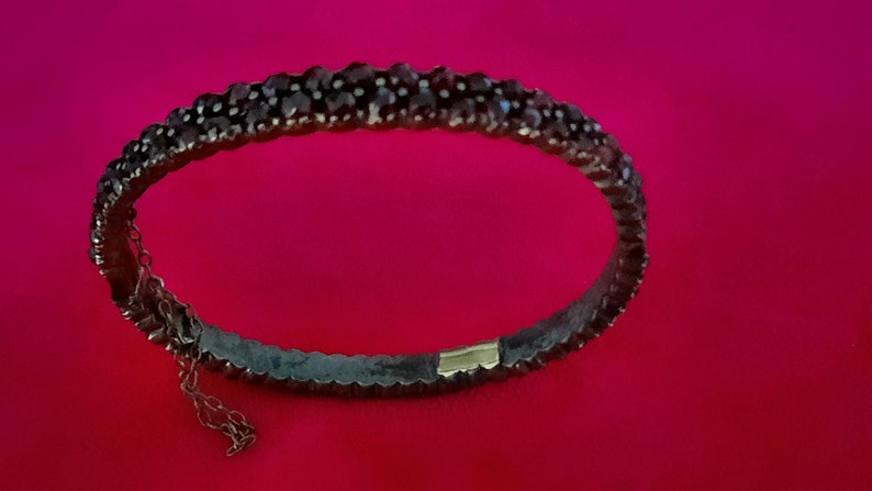 K&ouml;nnte beinhalten: Ein silbernes Armband mit einer Reihe dunkelroter Edelsteine. Das Armband hat einen Verschluss an der Seite.
