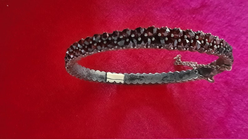 K&ouml;nnte beinhalten: Ein silbernes Armband mit einer Reihe dunkelroter Granatsteine. Das Armband hat einen Scharnierverschluss.