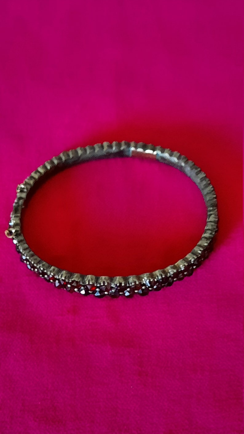 K&ouml;nnte beinhalten: Ein silbernes Armreif-Armband mit einer Reihe dunkelroter Edelsteine.