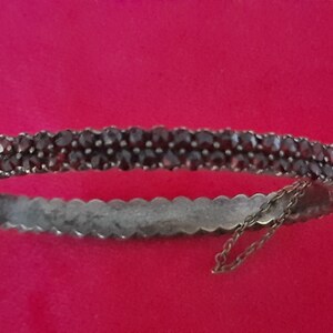 K&ouml;nnte beinhalten: Ein silbernes Armreif-Armband mit einer doppelten Reihe dunkelroter Granatsteine. Das Armband hat eine zarte Kette, die am Verschluss befestigt ist.