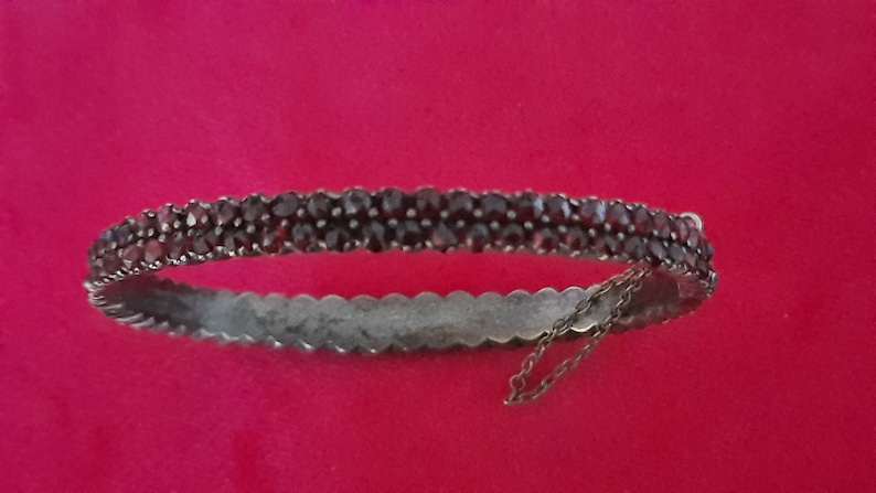 K&ouml;nnte beinhalten: Ein silbernes Armreif-Armband mit einer doppelten Reihe dunkelroter Granatsteine. Das Armband hat eine zarte Kette, die am Verschluss befestigt ist.