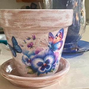 May include: A decorative, distressed-finish flower pot with a matching saucer. The pot is adorned with images of blue butterflies and pansies. The pot is made of a light brown material with a white wash finish.