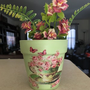 May include: A light green ceramic flower pot with a vintage teacup and roses design. The pot is decorated with pink and white flowers and butterflies.