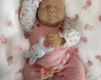Può includere: Bambola realistica che indossa un pagliaccetto rosa lavorato a maglia con bottoni in legno e maniche a fantasia floreale. La bambola tiene in mano un giocattolo a forma di coniglietto all'uncinetto e ha un ciuccio. La bambola è su una coperta bianca con stampe di coniglietti rosa.