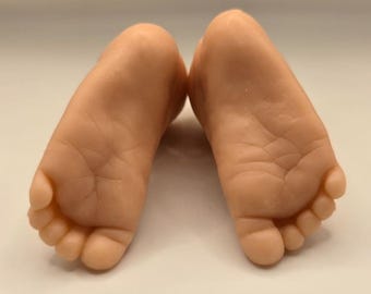 Può includere: Un paio di piedi di bambino realistici, color carne. I piedi sono posizionati fianco a fianco, con le piante rivolte verso l'alto. Le dita dei piedi sono ben definite e la pelle mostra dettagli fini.