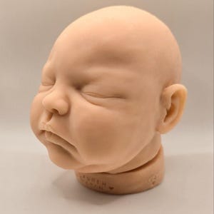 May include: A realistic, unpainted baby doll head with closed eyes and a neutral expression. The head is made of a light peach-colored material and is mounted on a round, tan-colored base. The base has text that reads "LAUREN TON".