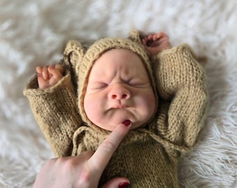 Può includere: Un neonato con un completo con cappuccio lavorato a maglia di colore marrone chiaro. Il viso del bambino è accigliato e un dito con lo smalto rosso tocca il mento del bambino. Il bambino è sdraiato su una superficie bianca e soffice.