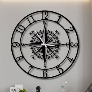Könnte beinhalten: Schwarze Kompassuhr aus Metall mit römischen Ziffern und einem Kompassrosen-Design. Die Uhr ist an einer weißen Wand in einem Wohnzimmer angebracht.
