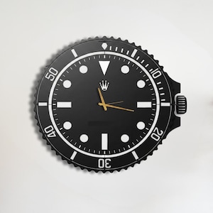 May include: A black wall clock with a white bezel and gold hands, resembling a watch face. The clock has the text "BLACK WALDECOR" in the upper right corner. The clock is mounted on a white wall above a wooden table with decorative items.