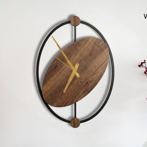 Pode incluir: Um relógio de parede moderno com um mostrador oval de madeira escura e ponteiros dourados, emoldurado em um círculo de metal preto. O relógio está montado em uma parede branca acima de uma mesa de madeira com itens decorativos.