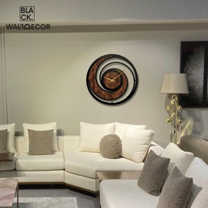 May include: A modern wall clock with a spiral design. The clock features a dark brown wooden center with a black outer frame. Gold-colored hands indicate the time. The text "BLACK. WALDECOR" is visible in the upper left corner.