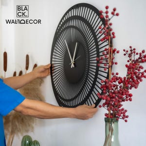 Side view of installing a large modern black metal wall clock on a white wall, showing lightweight design and easy mounting.
