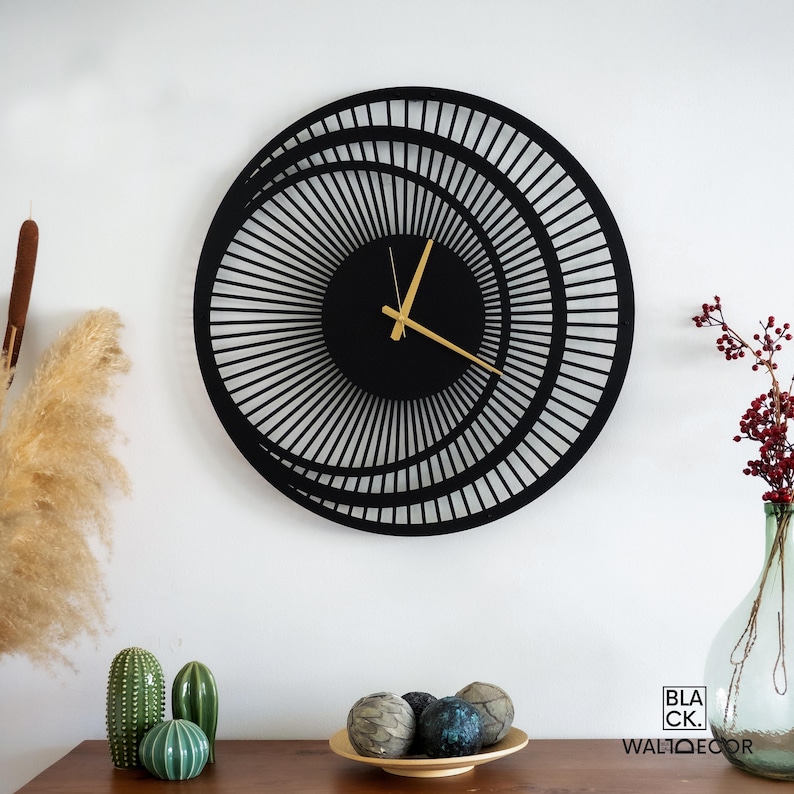 Modern black metal wall clock with gold hands styled above a wooden console with plants, minimalist living room decor.