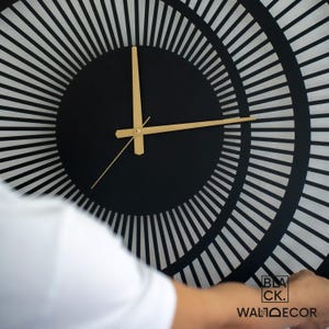 Close-up of modern black metal wall clock with gold hands and matte electrostatic finish, triple circle detailing.
