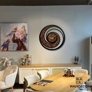 May include: A modern wall clock with a spiral design. The clock features a dark wood grain center, black metal frame, and gold-colored hands. The clock is mounted on a light gray wall, with a painting and furniture in the background.