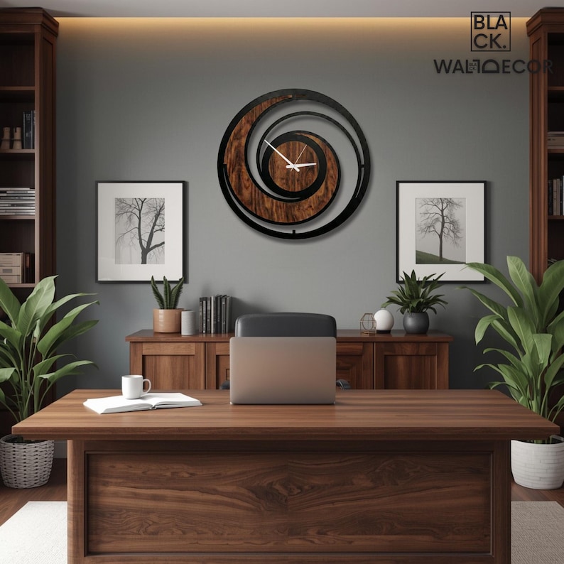 May include: A wooden desk with a laptop, a mug, and a notebook. A large, circular wall clock with a spiral design hangs on the gray wall. Two framed pictures of trees are on the wall. The clock has the text "BLACK. WALLDECOR".