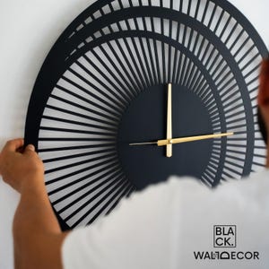 Man installing a large modern black metal wall clock on a white wall, demonstrating balanced 3D effect and easy hanging.