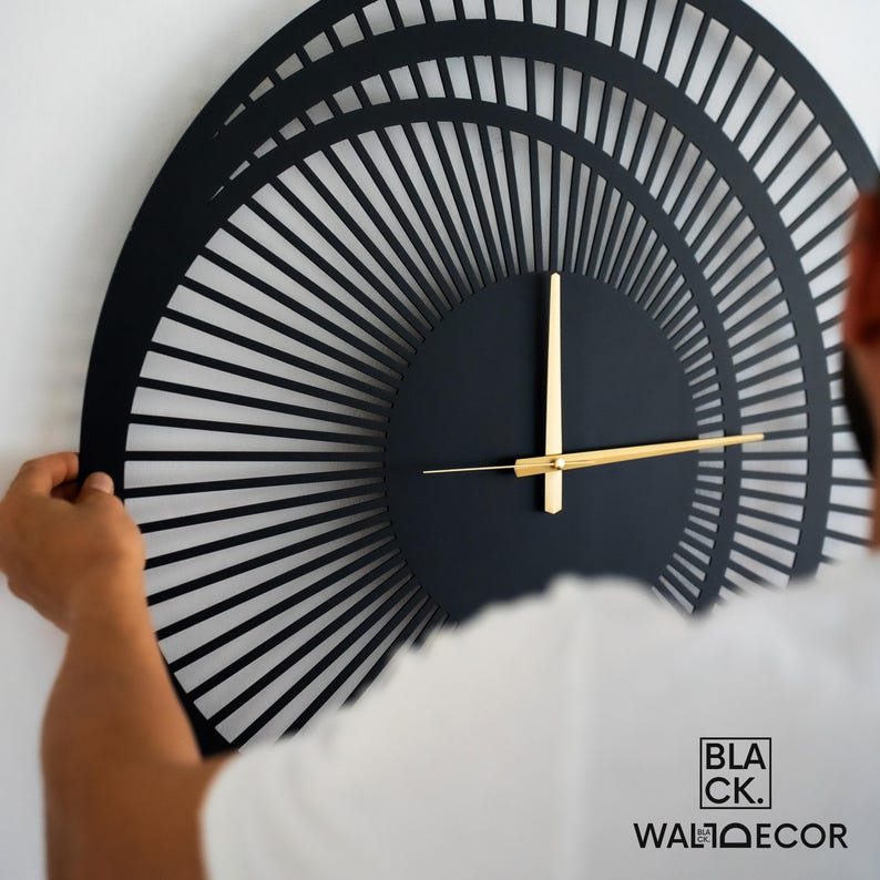 Man installing a large modern black metal wall clock on a white wall, demonstrating balanced 3D effect and easy hanging.