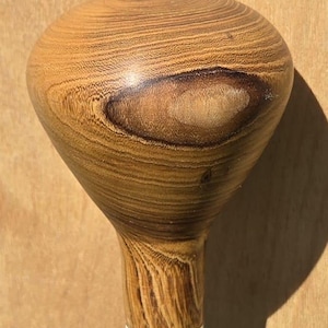 May include: A polished wooden handle with a rounded, bulbous top featuring prominent natural grain patterns and a dark, oval-shaped knot. The wood displays varying shades of light and medium brown, transitioning to a smooth, cylindrical base.