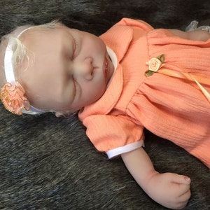 K&ouml;nnte beinhalten: Eine schlafende Reborn-Puppe, die ein pfirsichfarbenes Kleid mit wei&szlig;em Kragen und einem passenden Band tr&auml;gt. Die Puppe hat blondes Haar und tr&auml;gt ein wei&szlig;es Stirnband mit einer pfirsichfarbenen Blume.