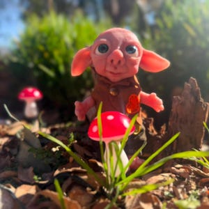 May include: A small, pink-skinned creature with large blue eyes and pointed ears, wearing a brown vest with a small orange heart-shaped detail. The figure stands among several red and white mushroom-shaped ornaments in a garden setting.