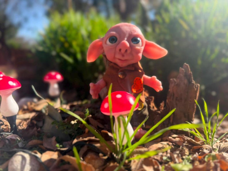 May include: A small, pink-skinned creature with large blue eyes and pointed ears, wearing a brown vest with a small orange heart-shaped detail. The figure stands among several red and white mushroom-shaped ornaments in a garden setting.
