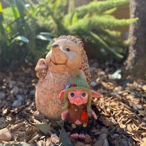 May include: A small, pink-skinned creature with large blue eyes, wearing a green knitted hat with a small red bead, a brown vest with a red heart patch, and dark trousers, standing in front of a textured, light brown stone hedgehog figurine.