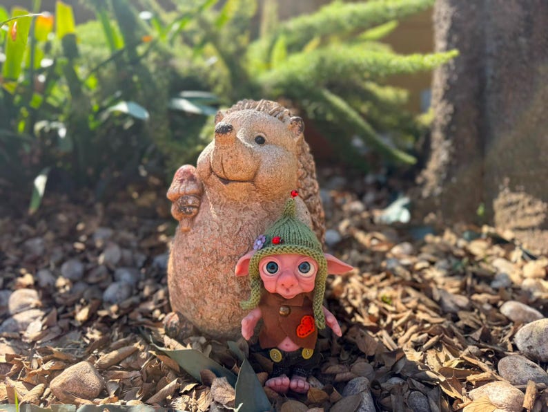 May include: A small, pink-skinned creature with large blue eyes, wearing a green knitted hat with a small red bead, a brown vest with a red heart patch, and dark trousers, standing in front of a textured, light brown stone hedgehog figurine.